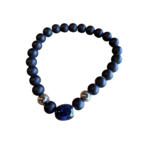 Bracelet en billes volcaniques /pierre bleue : comprend des perles volcaniques rondes de couleur noire mate, deux accents argentés brillants et une pierre ovale plus grande de couleur bleu foncé, sur fond blanc.
